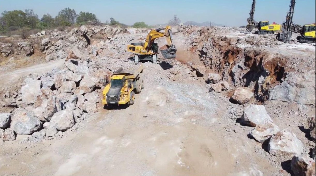 Excavator loading a dump truck in a quarry with drill rigs behind